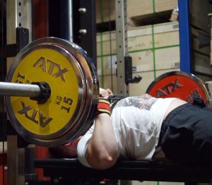 A person is performing a bench press in a gym. The barbell is loaded with large yellow and red weight plates labeled ATX. The individual is lying on a bench, focusing on lifting the weight. Boxes and shelving are visible in the background.