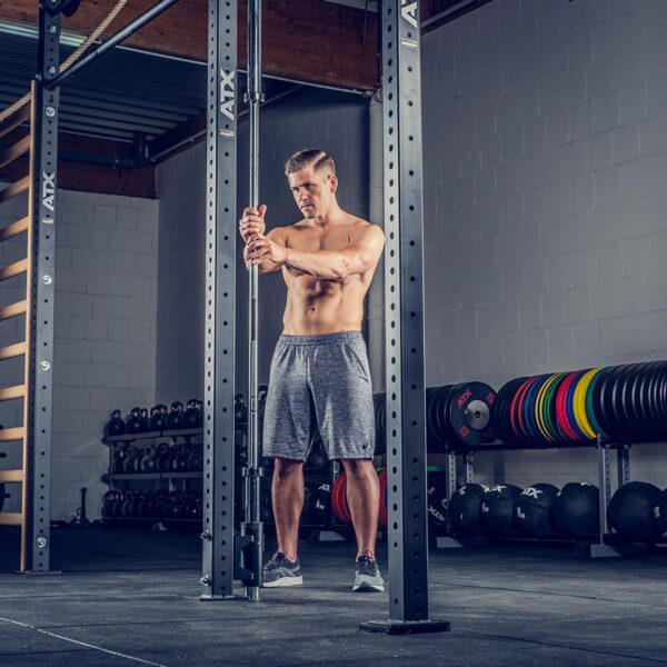 A shirtless man in gray shorts exercises with determination on a cable machine at a gym, equipped with kettlebells, weight plates, and a wooden ladder, alongside an ATX® Barbell Storage system.