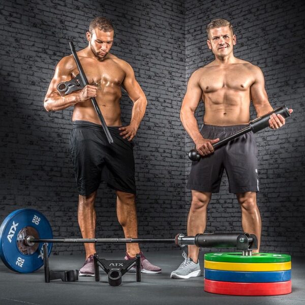 Two muscular men stand shirtless in a gym with an ATX® Post Landmine. One holds a rod, the other a barbell. Weight plates are stacked beside the barbell on the floor, all against a gray brick wall backdrop.