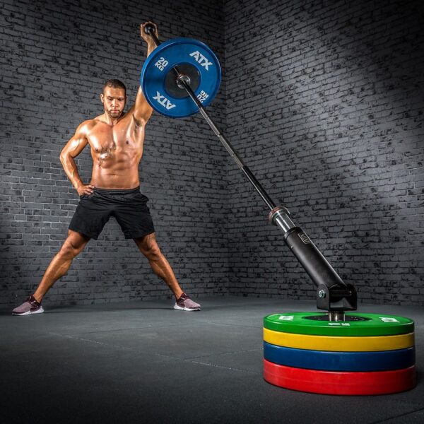 A muscular man is using the ATX® Post Landmine in the gym, gripping a barbell with one hand. Standing with legs wide apart against a brick wall, he demonstrates strength and precision while maneuvering the weights.