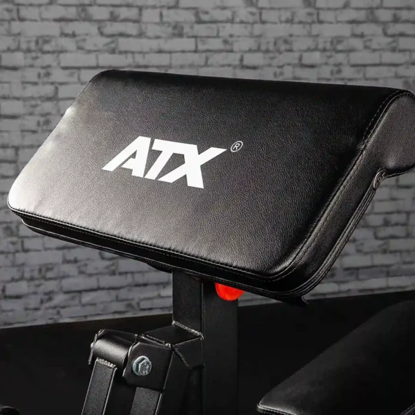 A close-up of a sleek black exercise bench featuring the ATX® logo, set against a backdrop of gray brick. Positioned near the ATX® Bicep Blaster Preacher Curl machine, it is part of a comprehensive gym or fitness equipment setup.