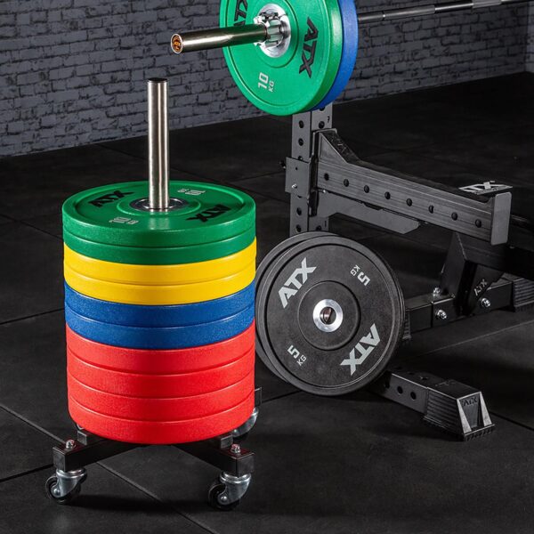 An ATX® Rolling Bumper Stacker holding colorful red, yellow, and green weights stands beside a gym bench. A barbell with extra weights is visible in the background, all set on a black rubber gym floor.