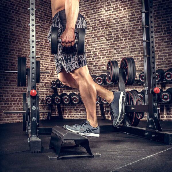 In front of brick walls and gym equipment, a person does step-ups on a low platform surrounded by racks of dumbbells. Holding two ATX® Calf Blocks, they showcase their strength and focus, dressed in athletic apparel and shoes.