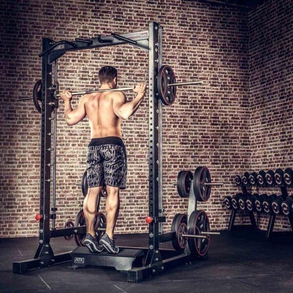A person performs pull-ups on a metal frame in a brick-walled gym, wearing shorts and sneakers. Nearby is an ATX® Calf Block, while weight plates are stored on racks in the background.