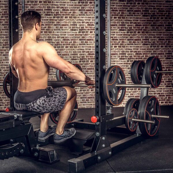 A person in shorts is seated on a gym bench performing a cable row, with the ATX® Calf Block complementing the equipment, including a weight rack with multiple plates, set against a brick wall backdrop.