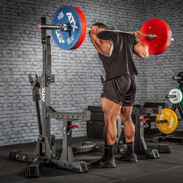 A person in black athletic clothing performs a squat with a heavily loaded barbell on an ATX® Competition Combo Rack in a gym. Weight plates are stacked on the floor nearby against a brick wall background.