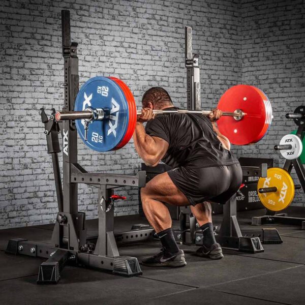 A person squats with a loaded barbell on their shoulders using the ATX® Competition Combo Rack. The barbell has red and blue weight plates, set against a gym backdrop featuring equipment and a brick wall.