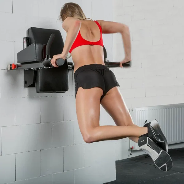 A person with a ponytail, dressed in a red sports bra, black shorts, and sneakers, is performing an upper body exercise on an ATX® Wall Mounted Knee Raise & Dip in a gym. A white brick wall and a radiator are visible in the background.