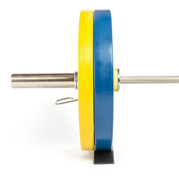 An ATX® Deadlift Wedge is positioned near a horizontally placed barbell, featuring yellow and blue weight plates secured by a metal clip. The bar rests on a black mat with its shiny metal rod extending to the right.