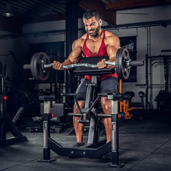A determined man in a red tank top and black shorts performs an intense workout with the ATX® EZ Bar Curl Camo - DISCONTINUED amid various gym equipment.