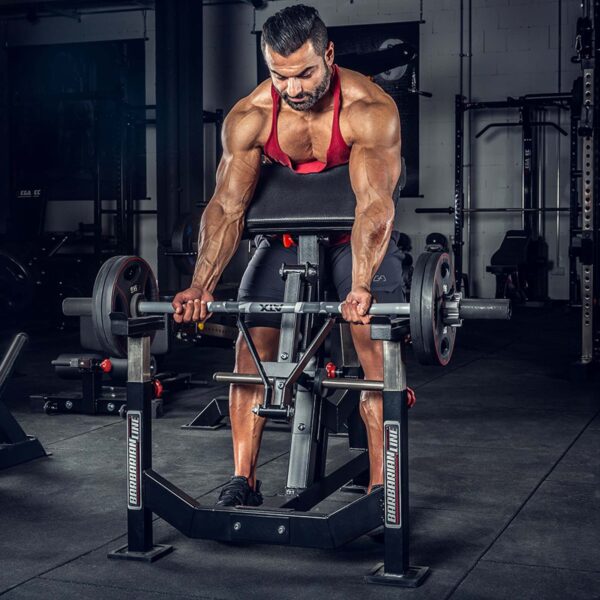 A muscular man in a dimly lit gym performs curls on a preacher bench using the now-discontinued ATX® EZ Bar Curl Camo. Hes dressed in a red tank top and black shorts, surrounded by various gym equipment.