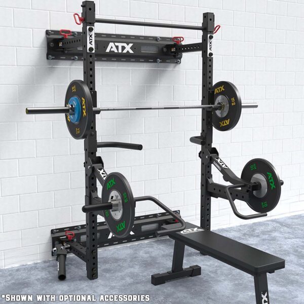 An ATX® Fold Back Rack Mount Rails with a loaded barbell on adjustable hooks, weight plates at the sides, and a bench underneath. Various gym accessories, including a weight holder and pull-up bar, are also visible.