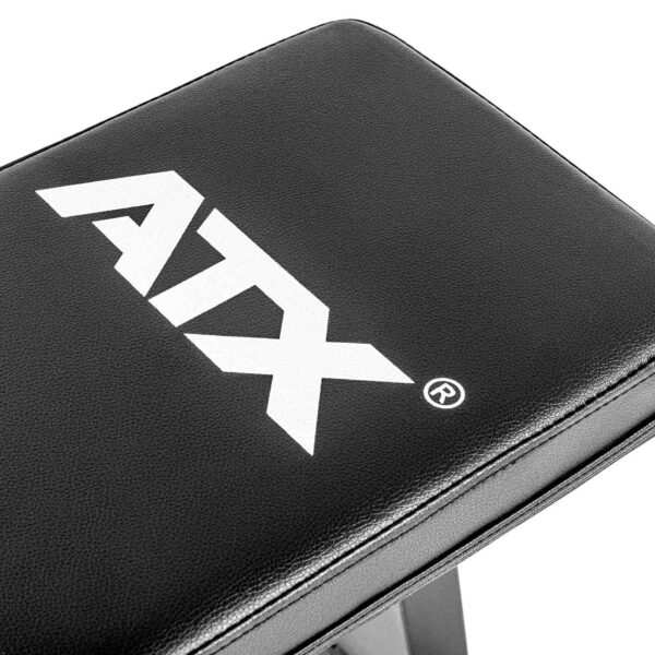 A close-up shows the ATX® Flat Bench Compact in black, featuring a white ATX logo. The exercise bench has a textured finish with visible stitching along the edges for enhanced durability and style.