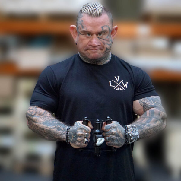 A person with tattoos and styled short hair is seen in a gym holding an ATX® Lat Bar Blackline, wearing a black t-shirt. The background shows blurred shelves.