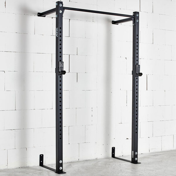 An ATX® Wall Mounted Squat Rack, black and featuring a pull-up bar, is installed on a white brick wall. It has adjustable J-hooks for securing a barbell. The concrete floor is clear of weights and other equipment.