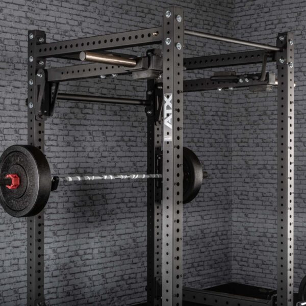 A black ATX® Jammer Arms power rack is set against a gray brick wall, featuring a loaded barbell with black weight plates. The rack includes multiple adjustment holes, displaying the ATX brand name on the side.
