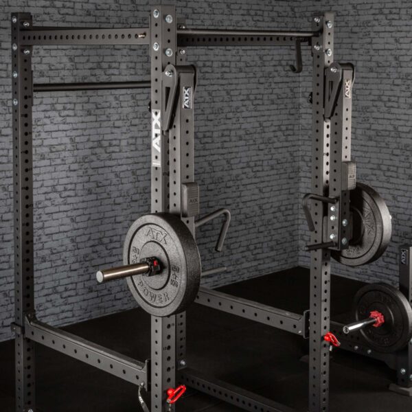 The black metal ATX® Jammer Arms power rack holds several weight plates on its side, set against a textured gray brick wall. It features various hooks and attachments for weightlifting.