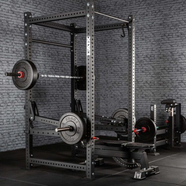 A home gym setup featuring ATX® Jammer Arms with a power rack, barbell, and weights on a dark floor against a gray brick wall. Additional fitness machines and weight plates can be seen in the background.