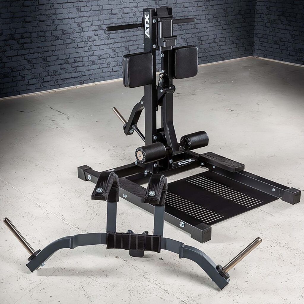 The ATX® Leg Master, a black and gray belt squat machine, is positioned in a gym with a textured gray brick wall and concrete floor. Equipped with padded supports, a platform, and handles for weightlifting, it expertly complements exercises like the standing leg curl.