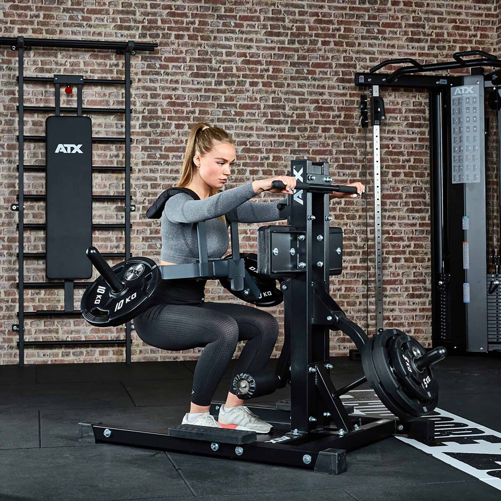 In a brick-walled gym, a person exercises on a seated calf raise machine with focus and determination, wearing workout gear. They are surrounded by various equipment, including the versatile ATX® Leg Master for comprehensive lower body strengthening.
