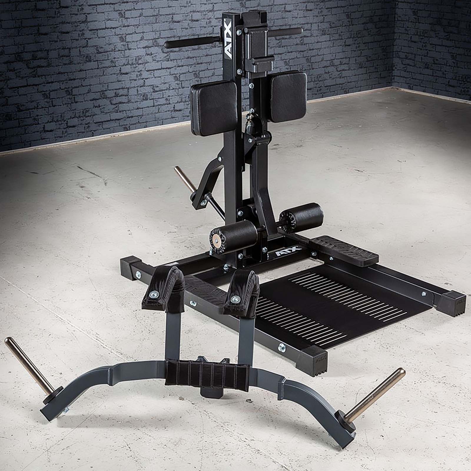 The ATX® Leg Master, a black and gray belt squat machine, is positioned in a gym with a textured gray brick wall and concrete floor. Equipped with padded supports, a platform, and handles for weightlifting, it expertly complements exercises like the standing leg curl.