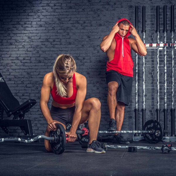 In a gym, a woman wearing a red sports top and black shorts kneels to adjust the weights on an ATX® Olympic Camo Power Bar. A man in a red sleeveless hoodie stands nearby, fixing his hood amid various gym equipment.