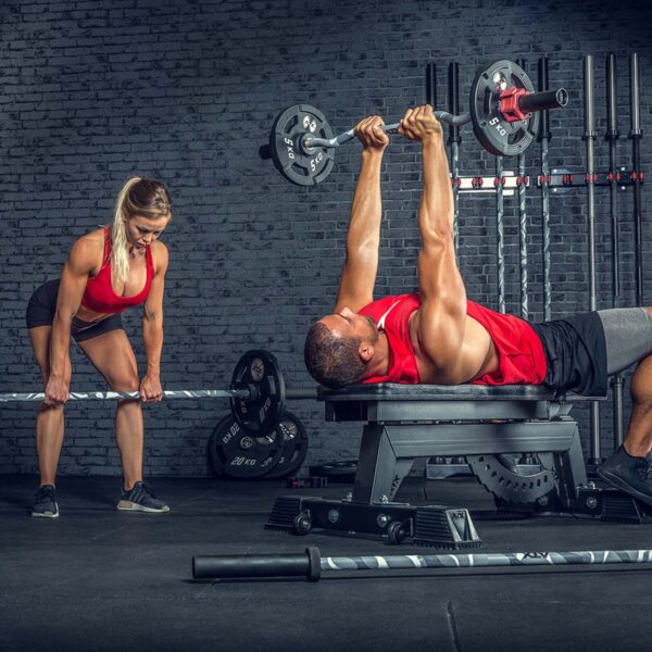 In a gym, one man on a bench is pressing an ATX® Olympic Camo Power Bar while a woman nearby holds another barbell. Both wear red tops, with various gym equipment lining the background wall.