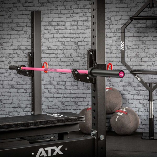 A gym setup includes a black squat rack with an ATX® Womens PINK 15kg Bar and an attached pull-up bar. Behind it, several ATX-branded medicine balls are stacked against a textured gray brick wall.