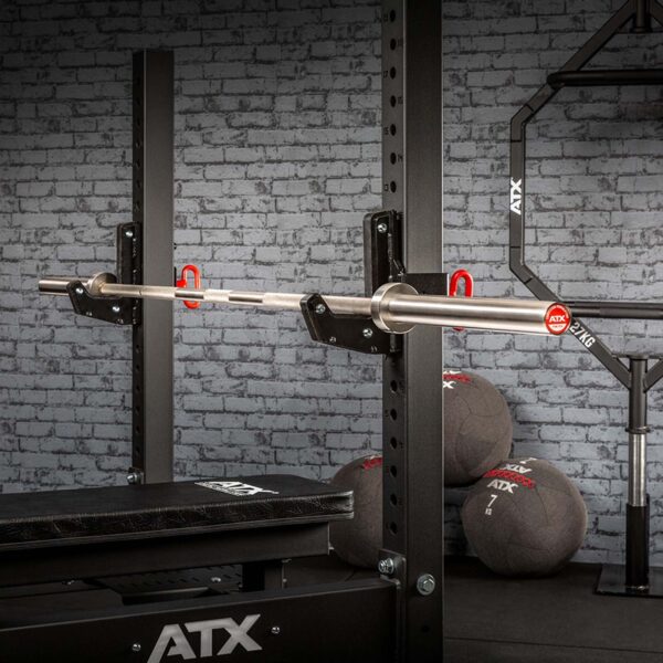 A gym setup features an ATX® Competition Powerlifting Barbell on a black squat rack against a gray brick wall. Three medicine balls are stacked in the background, with ATX prominently displayed on the equipment.