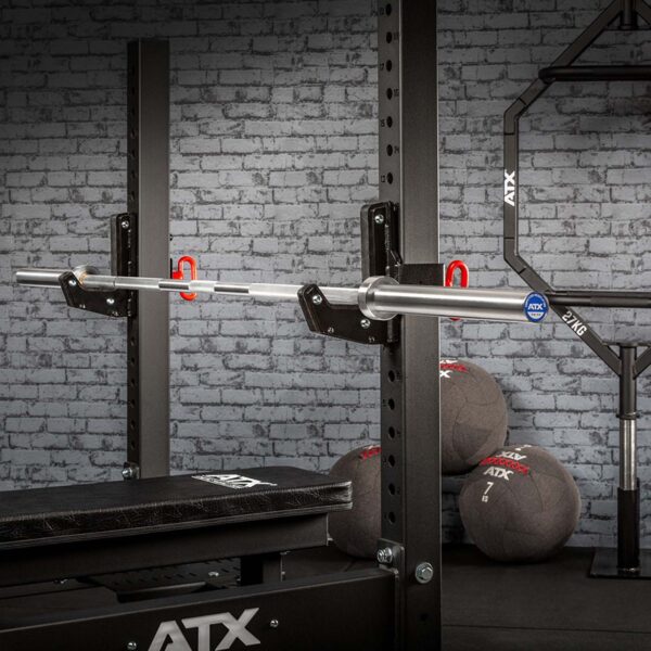 A gym setup includes an ATX® Competition Olympic Weightlifting Barbell on a rack with a bench below. Three medicine balls are stacked against a brick wall in the background, all branded with ATX.
