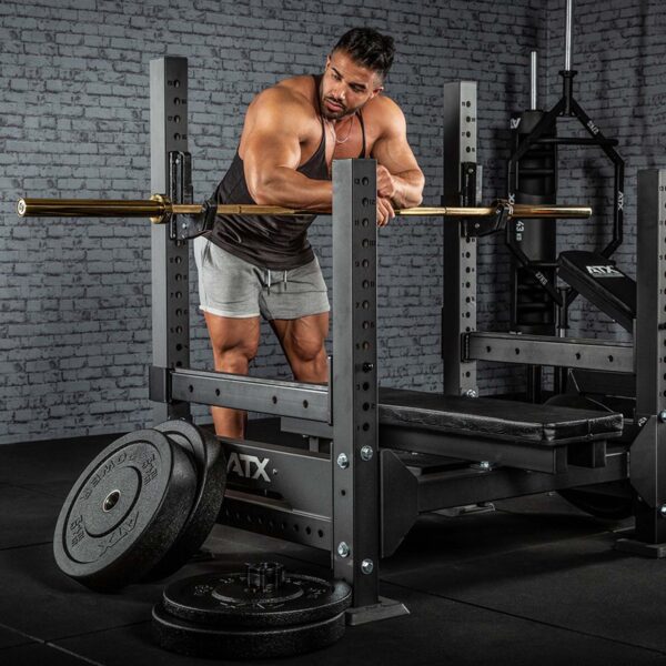 A man in a gym prepares a bench press by adjusting an ATX® Olympic Golden Powerlifting Bar on a squat rack. He wears a tank top and shorts, with weight plates on the floor nearby and brick walls visible in the background.