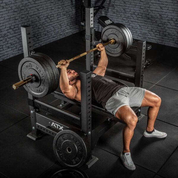 Using the ATX® Olympic Golden Powerlifting Bar, a person in a gym wears a dark tank top, gray shorts, and sneakers while bench pressing on a bench. The setting features dark flooring and a brick wall backdrop.