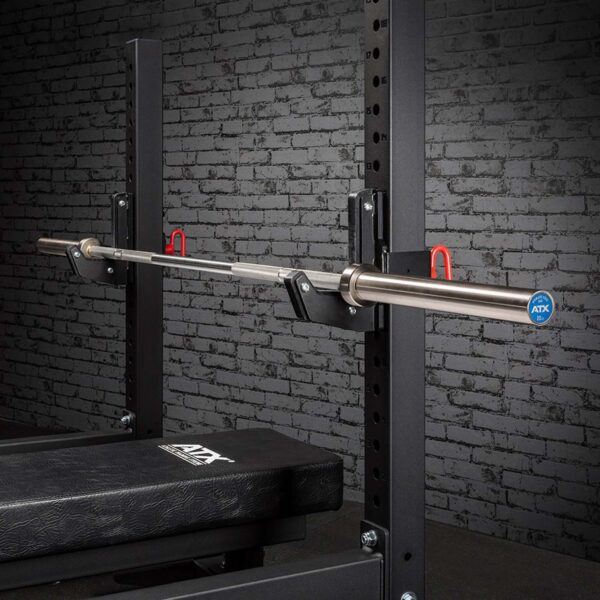 The ATX® Olympic Stainless Steel Bar is on a squat rack with safety hooks and a padded bench below, set against a textured gray brick wall, creating a gym environment.