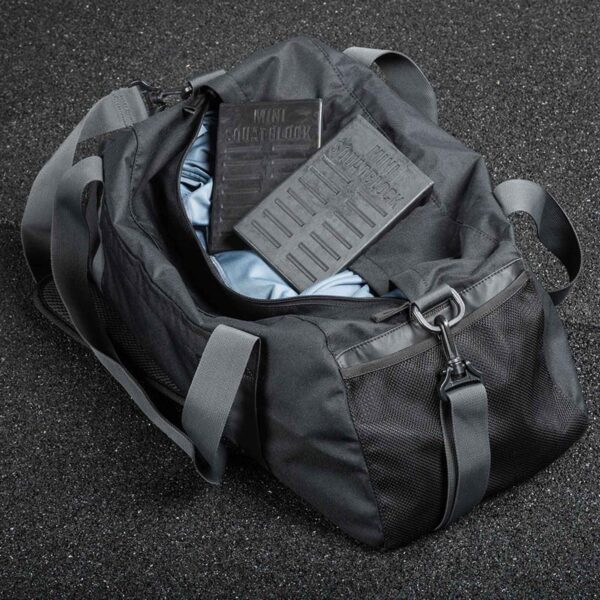 A black duffel bag is open on a speckled surface, showing a folded blue fabric and two rectangular black blocks labeled KNEE SLEEVE and SQUAT BLOCK. A mini squat wedge rests near gray straps and black mesh pockets.