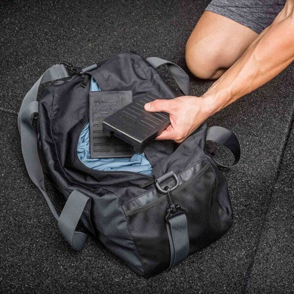 On a textured rubber floor, a person in gray athletic shorts kneels next to their trusty Mini Squat Wedges. They place a heavy weight plate into a black gym bag with teal interior lining and a wide strap for easy carrying.