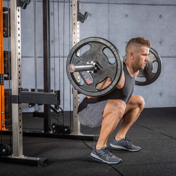 In the gym, a man expertly squats with a barbell on his shoulders. Clad in a black tank top, gray shorts, and sneakers, he employs Mini Squat Wedges to enhance his form on the padded floor near the squat rack.