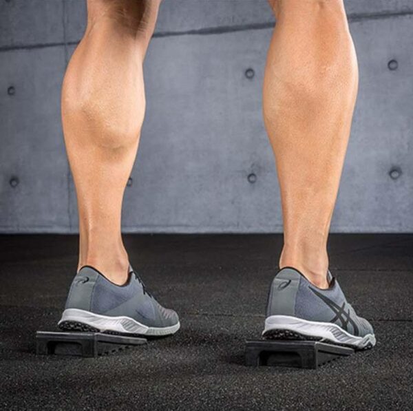 A person stands on Mini Squat Wedges wearing gray sneakers, accentuating their calf muscles against a minimalist gray wall and black floor.
