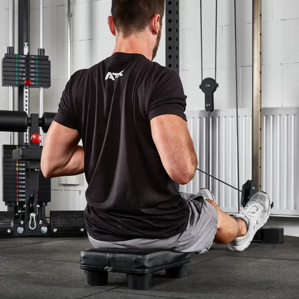 A man is seated on an ATX® Seated Row Seat in a gym, pulling cables. Hes wearing a black T-shirt, gray shorts, and white sneakers, with fitness equipment and weights visible in the background.