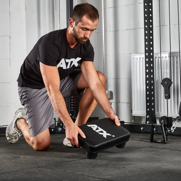 A man in a black ATX shirt and gray shorts kneels on a gym floor, placing an ATX® Seated Row Seat on the ground among various fitness equipment like a rack and weights.