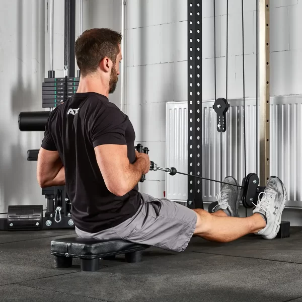 A person in a black shirt and gray shorts is sitting on the ATX® Seated Row Seat in a gym, pulling the cable toward their torso for a workout, with various gym equipment visible in the background.