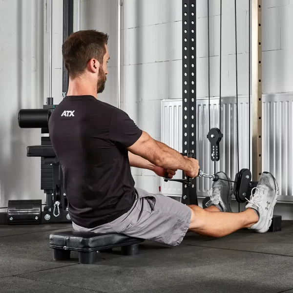 A person in a black ATX shirt and gray shorts is using the ATX® Seated Row Seat on a cable machine for rowing exercises. Theyre indoors, amidst gym equipment, facing away from the camera.
