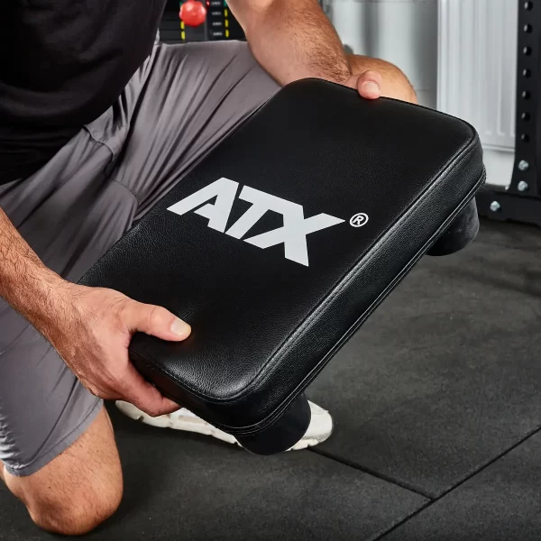 A person kneels on a gym floor, holding an ATX® Seated Row Seat with ATX written in white. Theyre wearing gray shorts and white sneakers, with fitness equipment visible in the background.