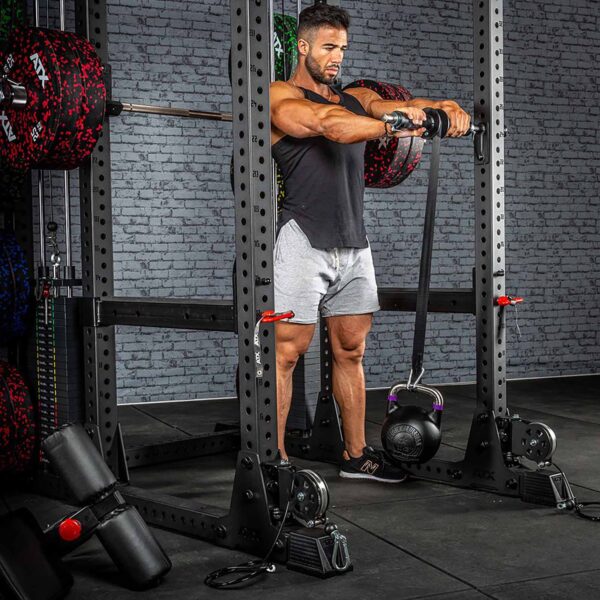 A man in a gym using the ATX® Universal Bracket during strength training with a barbell and kettlebell, stands inside a power rack, wearing a black sleeveless shirt and gray shorts amidst fitness equipment against a brick wall backdrop.
