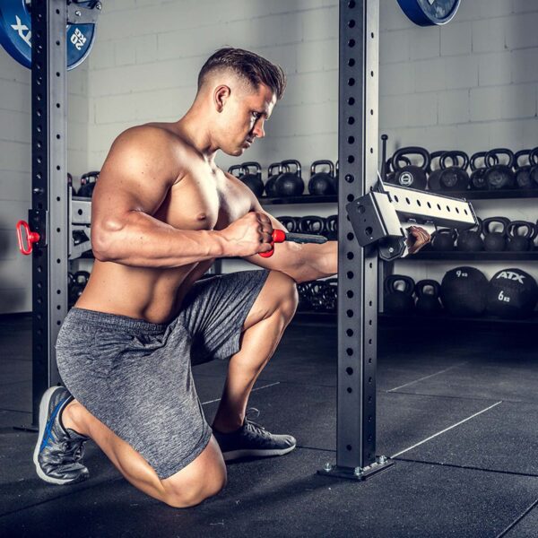 A muscular man kneels on the gym floor, focusing intently while adjusting ATX® Spotter Arms attached to a squat rack. He is shirtless, wearing gray shorts and sneakers, with kettlebells and weightlifting plates visible in the background.