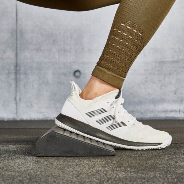 Close-up of a person wearing white athletic shoes with a gray sole, standing on a squat wedge block on the gym floor. Theyre sporting brown leggings with textured patterns, against a backdrop of a concrete wall.