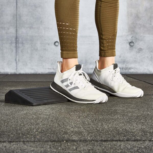 A person wearing white athletic shoes and brown leggings is using a squat wedge block for an exercise. The background is a gray, textured wall, and the floor is a dark, textured mat.