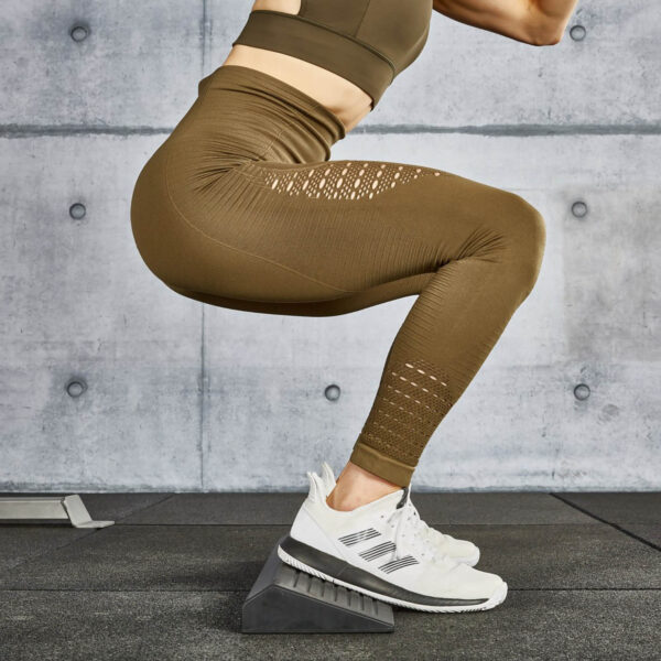 A person wearing brown leggings and white sneakers performs a squat on a squat wedge block in a gym setting. The wall in the background is made of concrete panels.