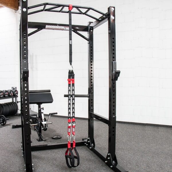 A gym setup with a black, sturdy power rack with adjustable safety bars and the ATX® Suspension Trainer Set PRO hanging from the top. An adjustable bench with black padding complements the scene, all set on a gray carpeted floor.
