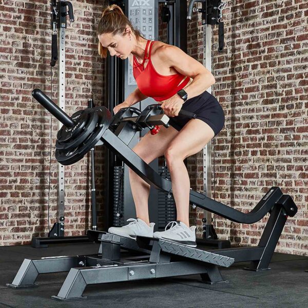 A woman in a red tank top and black shorts works out on an ATX T-Bar Row machine in a gym. She grips the handles and leans forward, focused on her training. The gym has a rustic brick wall and neatly arranged equipment.