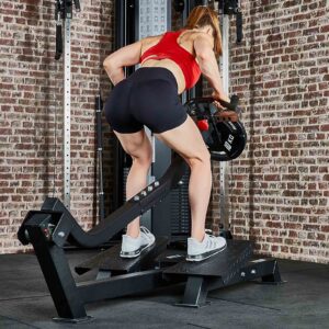 A person in a red top and black shorts performs a plate-loaded T-Bar Row with a 10 kg weight plate on an ATX gym machine. They are in a fitness area with a brick wall background.